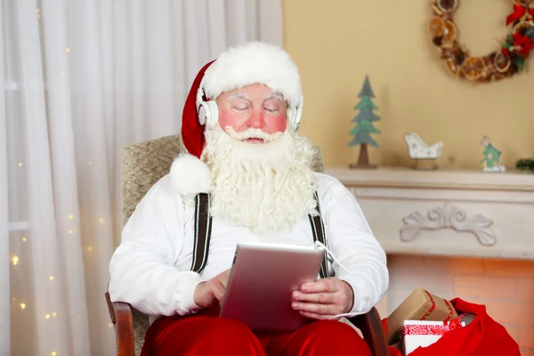 Santa claus zitten met digitale tablet in comfortabele stoel in de buurt van open haard thuis — Stockfoto