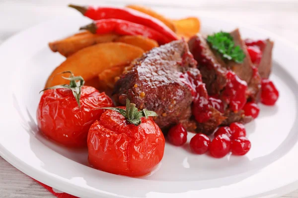 Вкусное жареное мясо с клюквенным соусом и жареными овощами на тарелке, на фоне цвета дерева — стоковое фото