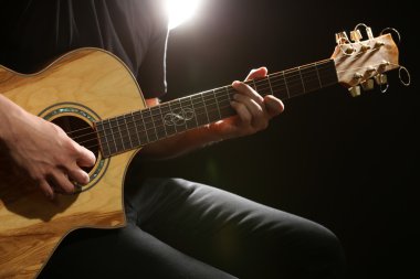 Genç adam akustik gitarla karanlık arka planda çalıyor.