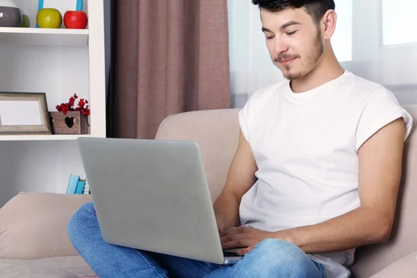 Hombre joven guapo sentado en el sofá y el uso de la computadora portátil en la habitación —  Fotos de Stock