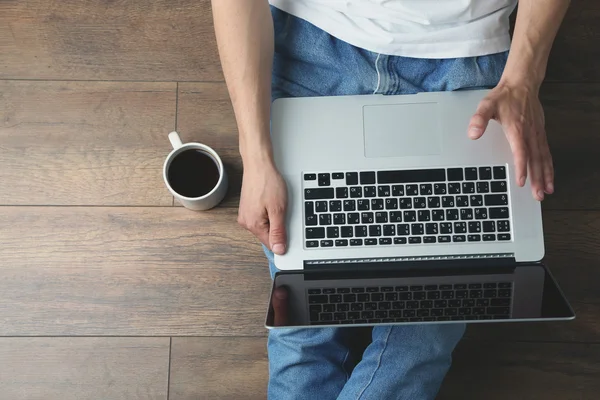 Hombre joven sentado en el suelo con ordenador portátil y taza de café en la habitación — Foto de Stock