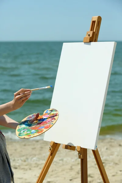 Female hand holding palette with paints and easel with canvas on beach — Stock Photo, Image