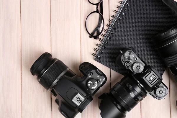 Modern cameras with glasses and notebook on wooden table, top view — Stock Photo, Image