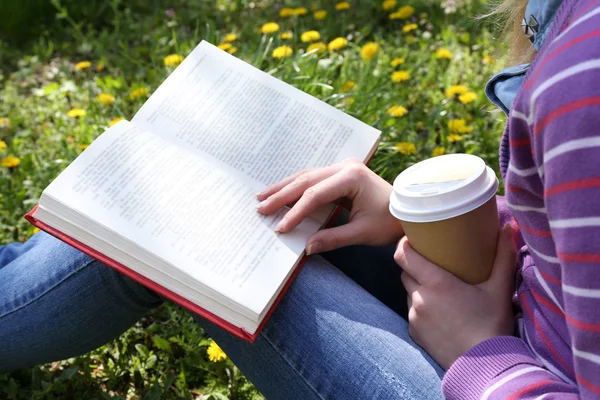 Молода жінка з книгою і чашкою кави сидить на зеленій траві на відкритому повітрі — стокове фото