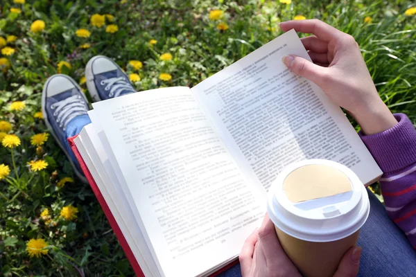 Giovane donna con libro e tazza di caffè seduta su erba verde all'aperto — Foto Stock