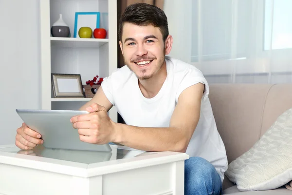 Hombre joven guapo sentado en el sofá y el uso de la tableta en la habitación —  Fotos de Stock