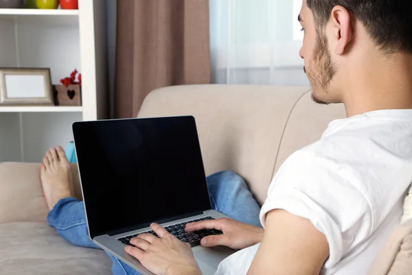 Hombre joven guapo sentado en el sofá y el uso de la computadora portátil en la habitación — Foto de Stock