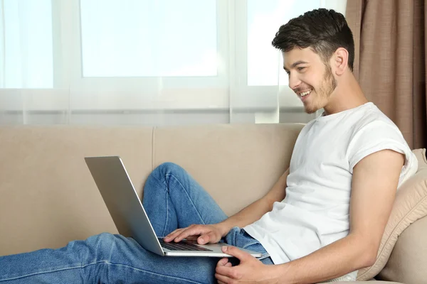 Hombre joven guapo sentado en el sofá y el uso de la computadora portátil en la habitación —  Fotos de Stock