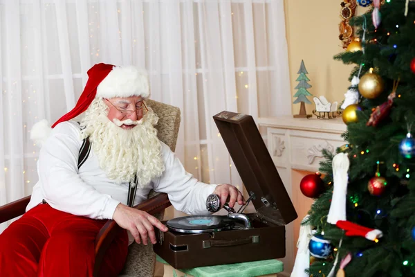 Weihnachtsmann sitzt in bequemem Stuhl in der Nähe von Retro-Plattenspieler zu Hause — Stockfoto