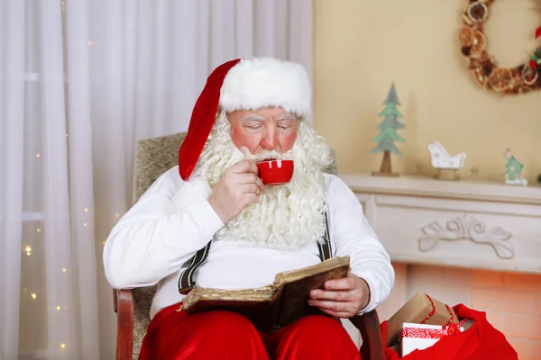 Santa claus zitten met een boek in comfortabele stoel in de buurt van open haard thuis — Stockfoto