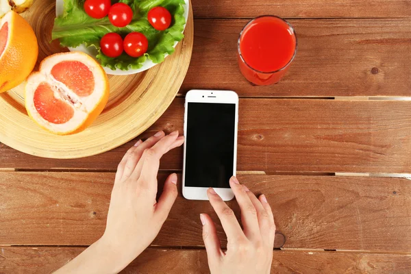 Food and mobile phone in female hands on wooden background — Stock Photo, Image