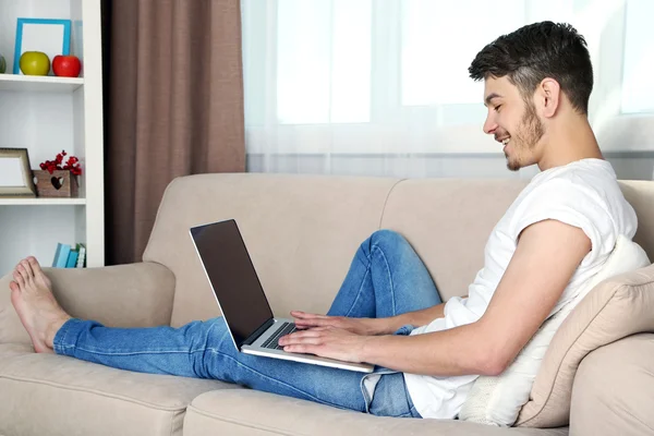 Hombre joven guapo sentado en el sofá y el uso de la computadora portátil en la habitación — Foto de Stock