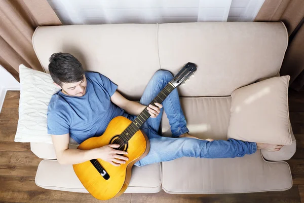 Молодой человек с гитарой на диване в комнате — стоковое фото