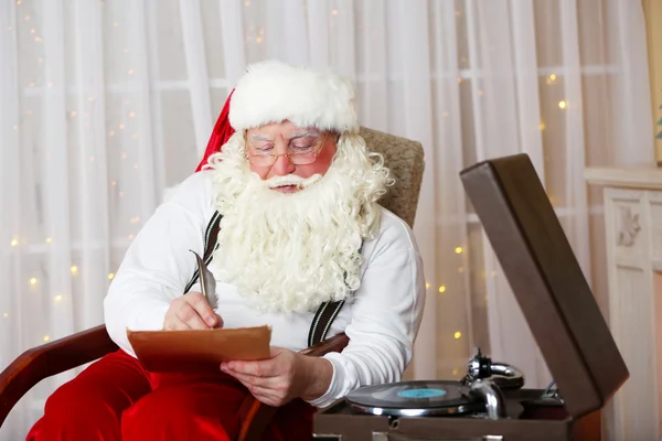 Weihnachtsmann sitzt mit Wunschzettel für Kinder im bequemen Stuhl am heimischen Kamin — Stockfoto