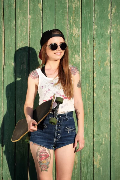 Gyönyörű lány tetovált test, gördeszka, fából készült fal háttér gazdaság — Stock Fotó