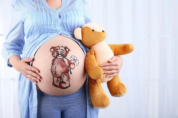 Piękna młoda kobieta w ciąży z zabawkami dziecka i obraz na brzuchu, na jasnym tle — Zdjęcie stockowe