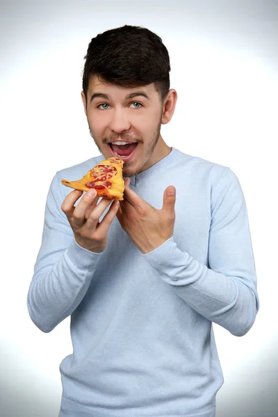 Junger Mann isst Stück Pizza auf grauem Hintergrund — Stockfoto