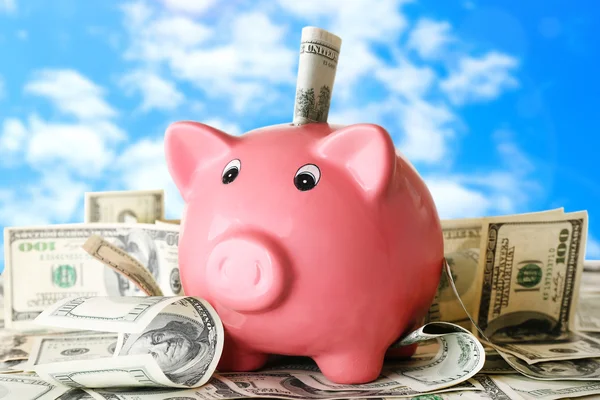 Piggy bank on pile of dollars on sky background — ストック写真