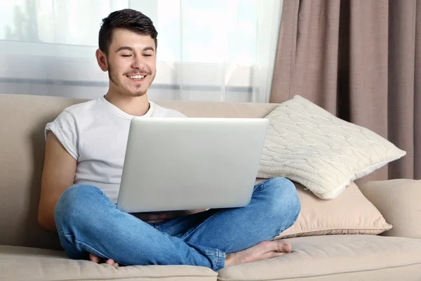 Bonito jovem sentado no sofá e usando laptop no quarto — Fotografia de Stock