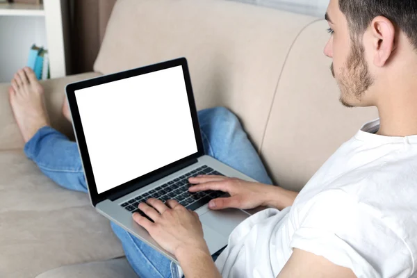 Schöner junger Mann sitzt auf Sofa und benutzt Laptop im Zimmer — Stockfoto