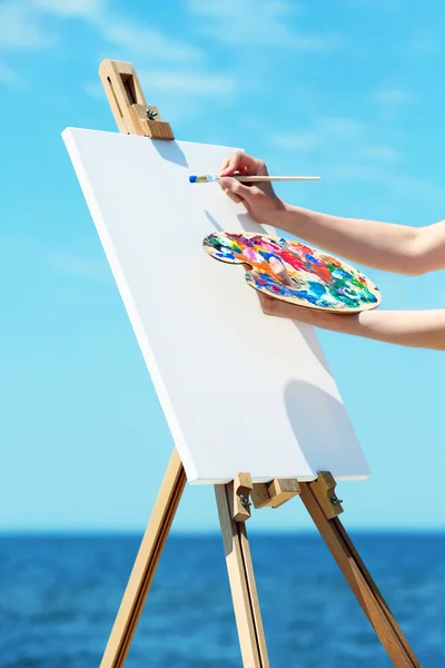 Female hand holding palette with paints and easel with canvas on beach — Stock Photo, Image