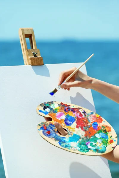 Female hand holding palette with paints and easel with canvas on beach — Stock Photo, Image