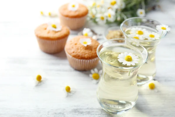 Bicchieri di tè alla camomilla con fiori di camomilla e gustosi muffin su sfondo di legno di colore — Foto Stock