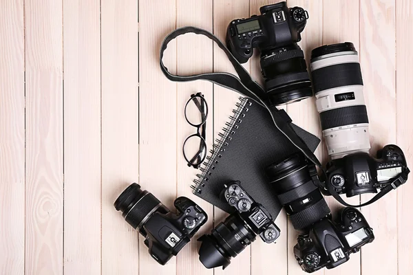 Modern cameras with glasses and notebook on wooden table, top view — Stock Photo, Image