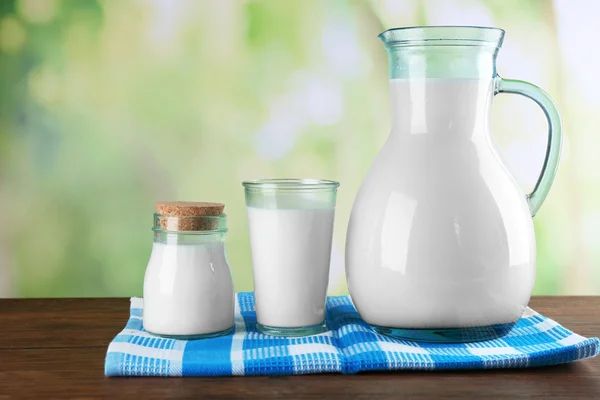 Brocca, barattolo e bicchiere di latte sul tavolo di legno, sullo sfondo della natura — Foto Stock