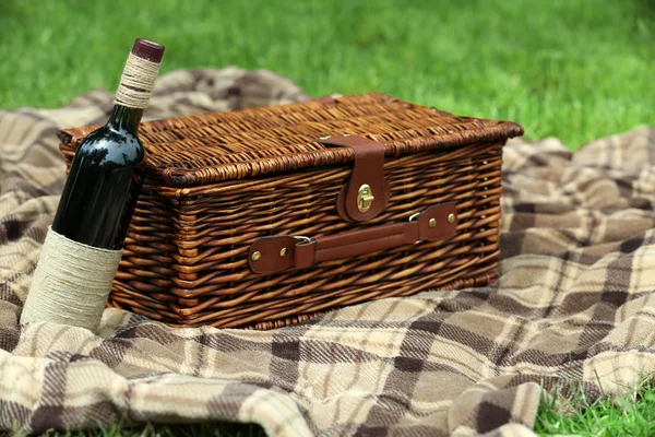 Wiklinowy kosz piknikowy, butelka wina i pled na zielonej trawie, na zewnątrz — Zdjęcie stockowe