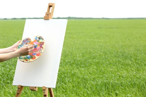 Female hand holding palette and easel with canvas in green field — Stock Photo, Image
