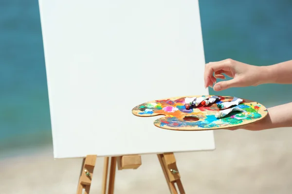 Female hand holding palette with paints and easel with canvas on beach — Stock Photo, Image