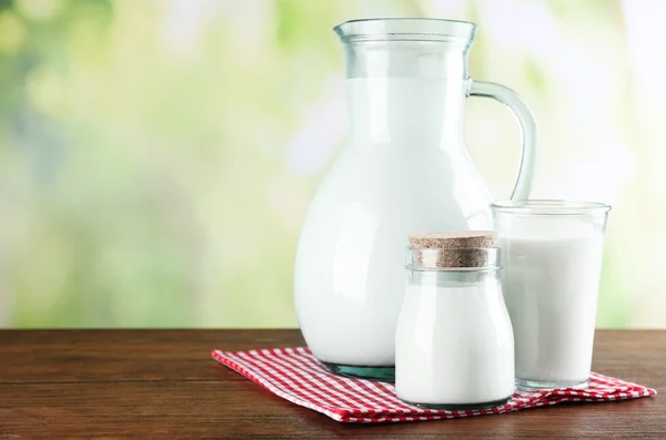 Pitcher, jar and glass of milk on wooden table, on nature background — Stock Photo, Image