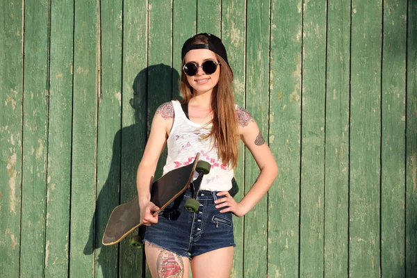 Menina bonita com corpo tatuado, segurando skate no fundo da parede de madeira — Fotografia de Stock