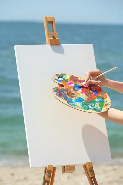 Mão feminina segurando paleta com tintas e cavalete com tela na praia — Fotografia de Stock