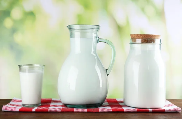 Pitcher, jar and glass of milk on wooden table, on nature background — Stock Photo, Image