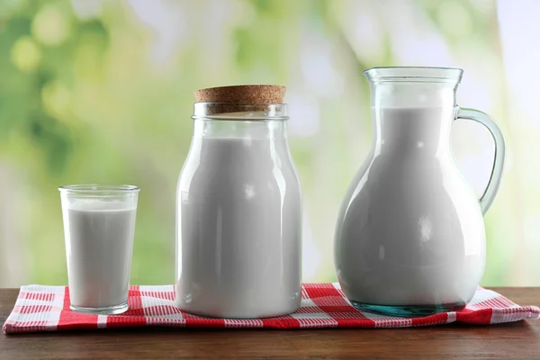 Pitcher, jar and glass of milk on wooden table, on nature background — Stock Photo, Image