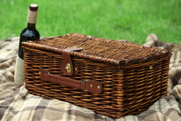 Wicker picnic basket, wine bottle and plaid on green grass, outdoors — Stock Photo, Image