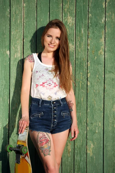 Όμορφο κορίτσι με τατουάζ σώμα, κρατώντας skateboard σε φόντο ξύλινα τοίχων — Φωτογραφία Αρχείου
