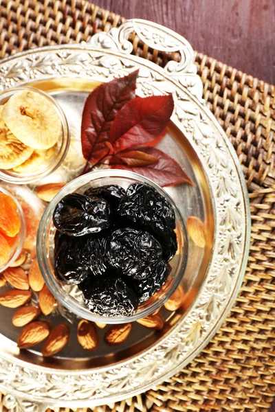 Pruneaux et autres fruits secs dans des soucoupes en verre avec feuilles de raisin sur plateau en métal et tapis en osier, vue de dessus — Photo