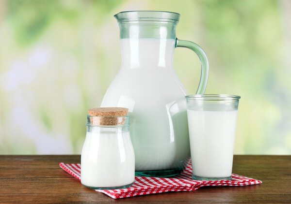 Pitcher, jar and glass of milk on wooden table, on nature background