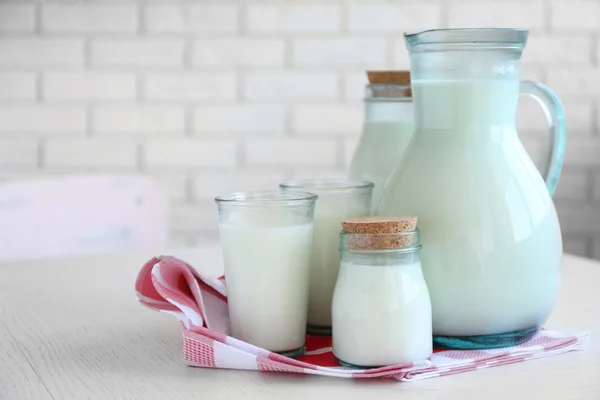 Jarra, frascos y vaso de leche sobre mesa de madera, sobre fondo de pared de ladrillos — Foto de Stock