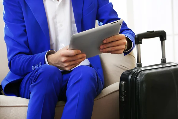 Business man with suitcase and tablet sitting on sofa in hall — Stock Photo, Image