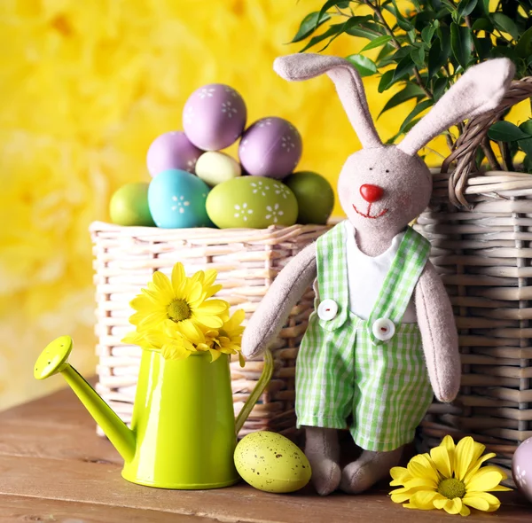 Пасхальный кролик с раскрашенными пасхальными яйцами с цветами на деревянном столе на желтом фоне — стоковое фото