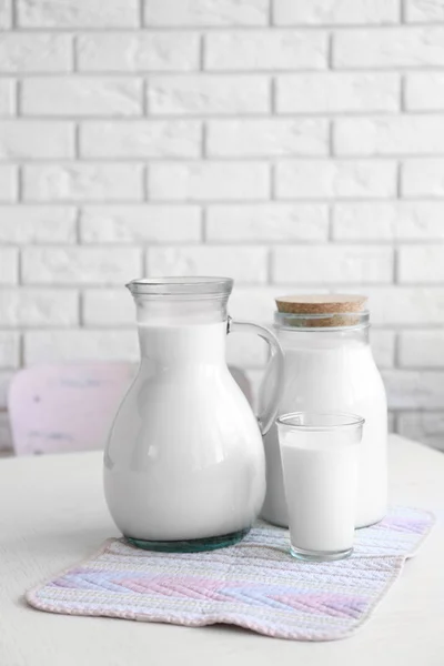 Pitcher, jar and glass of milk on wooden table, on bricks wall background — Stock Photo, Image