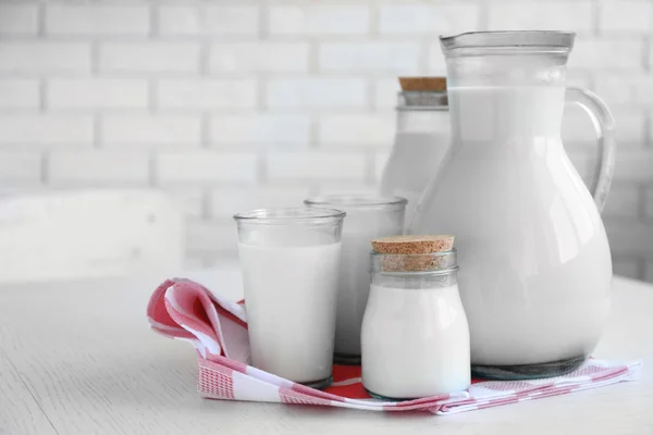 Pitcher, jars and glass of milk on wooden table, on bricks wall background — Stock Photo, Image