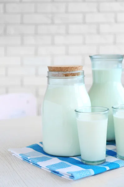 Pitcher, jar and glass of milk on wooden table, on bricks wall background — Stock Photo, Image