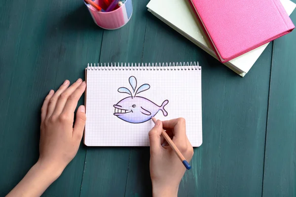 Mains féminines dessin baleine dans un cahier sur fond de table en bois — Photo