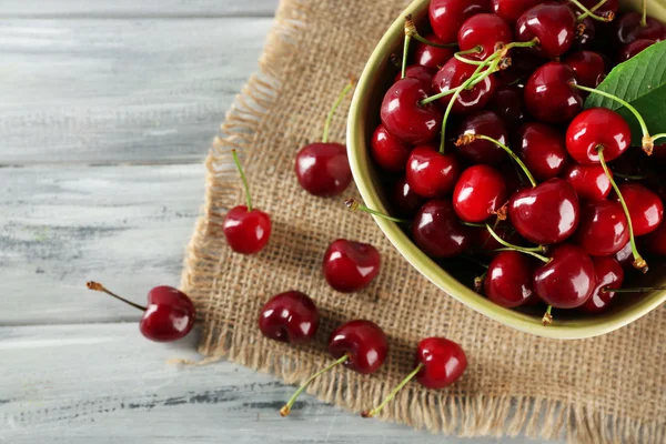 Cerezas frescas en tazón con saco sobre mesa de madera, primer plano — Foto de Stock