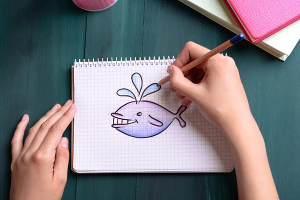 Manos femeninas dibujando ballena en cuaderno sobre fondo de mesa de madera —  Fotos de Stock